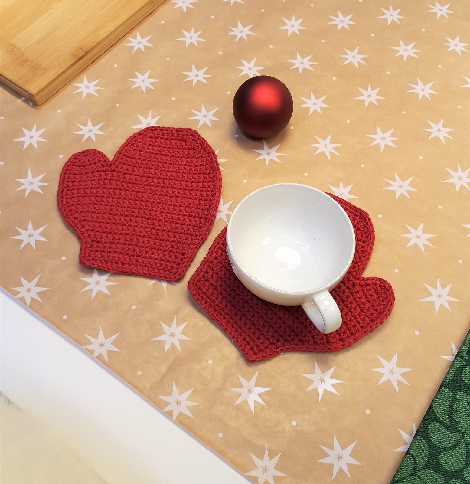 Two red crochet mitten coasters, one under a mug, with a red bauble on festive star-print paper