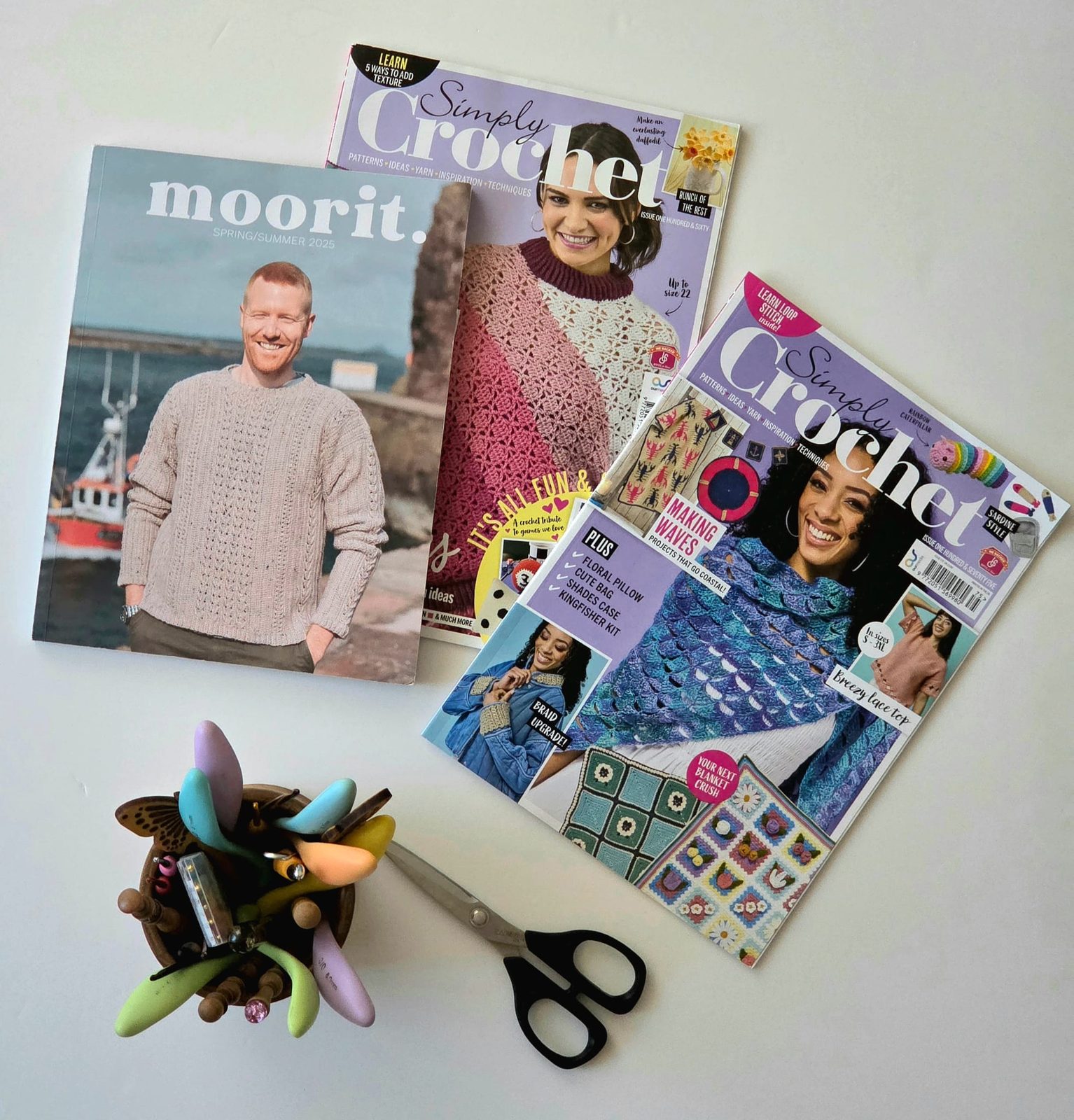 Moorit Spring/Summer 2025 and two issues of Simply Crochet fanned out on a table beside a pot of crochet hooks and a pair of scissors — the three covers carrying Asya's designs